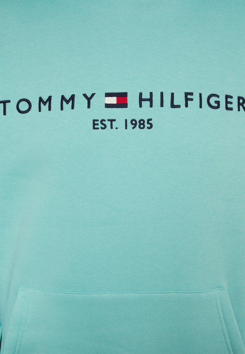 Tissu bleu clair avec texte brodé "Tommy Hilfiger" et logo en rouge, blanc et bleu, avec "EST. 1985" sous le logo.