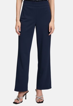 Woman wearing high-waisted navy wide-leg pants and black high-heeled sandals with crisscross silver straps against a plain background.