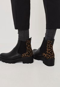 Černé kotníkové boty s leopardím potiskem, elastickými postranními panely, kulatou špičkou a hrubou texturou podrážky. Nošeny s šedými žebrovanými ponožkami a černými kalhotami.