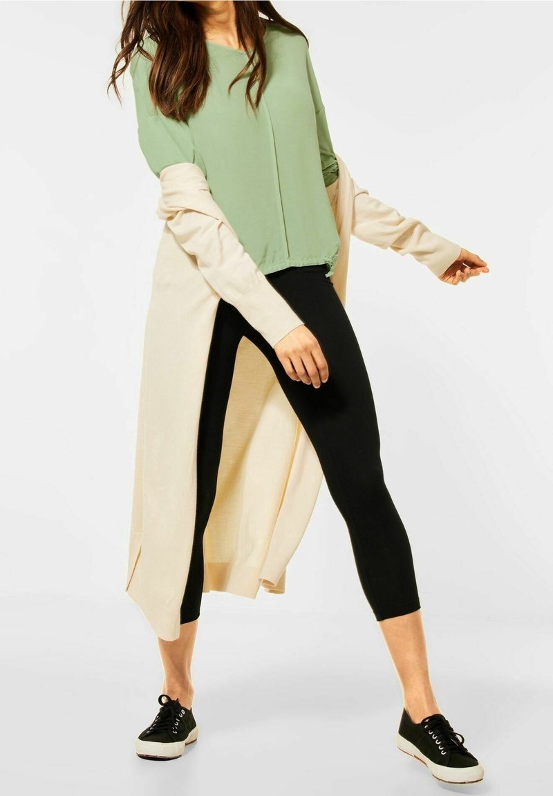 Green short-sleeve top layered with a light beige cardigan, paired with black leggings and black sneakers with white soles.