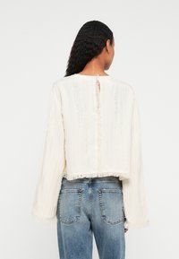 Frau mit langen, lockigen Haaren, die einen lockeren weißen strukturierten Pullover mit fransigen Kanten und hellblauen Jeans trägt, mit dem Rücken zur Kamera vor einem weißen Hintergrund.