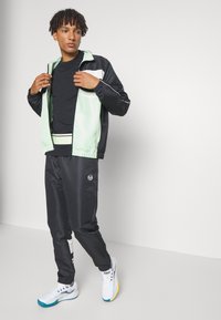 Juodos ir mėtų žalios spalvos sportinis treningas, turintis užtrauktuką ir elastingas rankoves, derinamas su atitinkamomis kelnėmis. Baltos sportinės bateliai su mėtų akcentais.