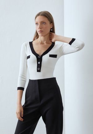 Bela rebrasta bodysuit z črnimi detajli, ki ima V-izrez, tri gumbe in majhne žepke na prsih, kombinirana z visokimi črnimi hlačami.