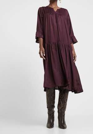 Kobieta w luźnej, długorękawowej purpurowej sukience midi w zestawieniu z brązowymi wysokimi kozakami na obcasie z wzorem skóry węża.