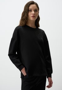 Felpa nera a maniche lunghe con scollo rotondo e vestibilità rilassata, caratterizzata da rhinestones decorativi disposti in un motivo a spirale su un lato.