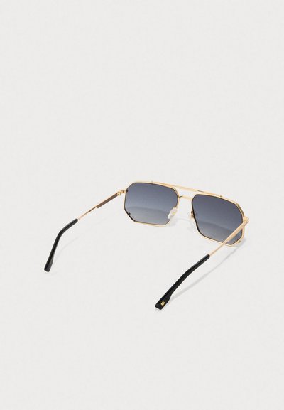 Gafas de sol octogonales doradas con lentes gris degradado. Montura metálica delgada, puntas de las patillas negras y un sutil detalle del logo en el brazo.