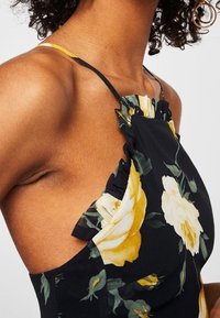 Robe florale en tissu noir avec des motifs de roses jaunes et blanches, décolleté froncé et fines bretelles, mettant en valeur une silhouette ajustée.