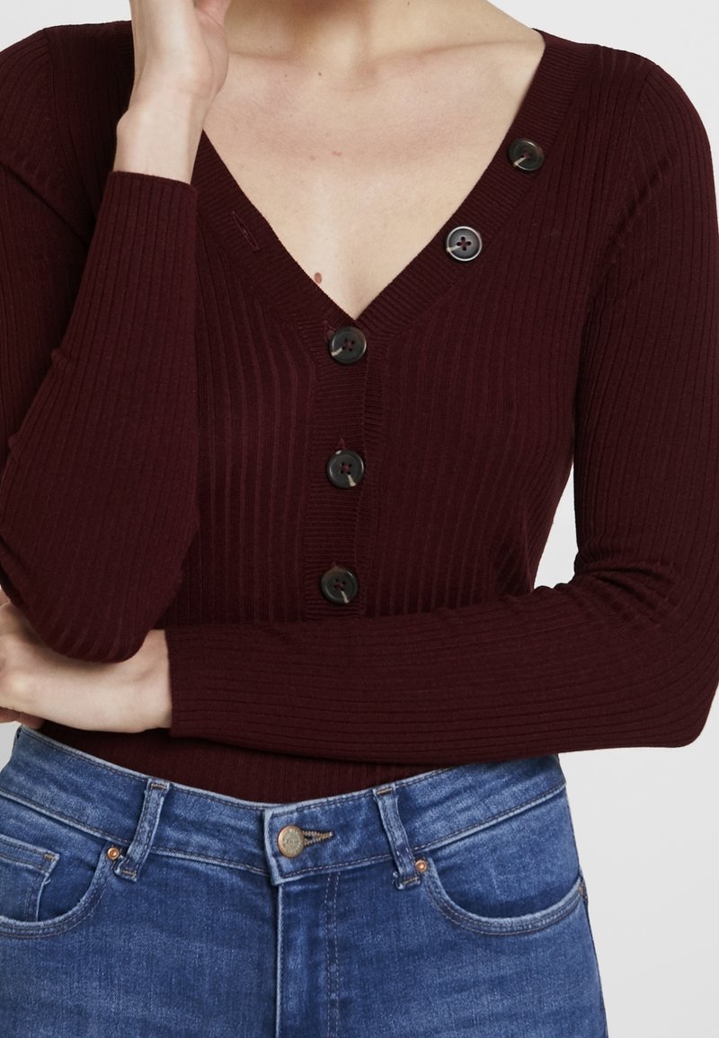 Femme portant un cardigan côtelé grenat avec des boutons noirs et un jean bleu, bras croisés, main touchant légèrement le menton.