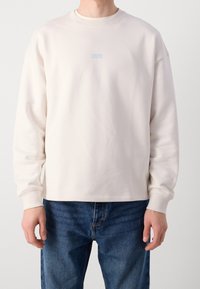 Uomo che indossa un semplice maglione a maniche lunghe color beige chiaro con un piccolo logo blu "HUGO" sul petto e jeans blu.