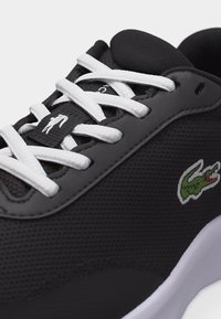 Chaussure de sport noire avec lacets blancs, tige en mesh, semelle texturée et logo crocodile vert sur le côté.