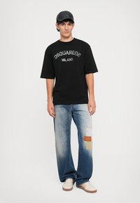 T-shirt nera in cotone con testo "DSQUARED2 MILANO", abbinata a jeans blu chiaro con dettagli in patchwork e sneakers crema. Cappellino grigio.