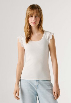 Młoda kobieta o prostych, jasnobrązowych włosach, ubrana w biały, bezrękawnik z koronkowymi rękawami z ażurowym wzorem oraz jasnoniebieskie dżinsy na jednolitym tle.