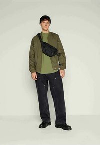 Olive green bomber jacket, matching tee, black fanny pack, loose dark pants with pockets, and black boots. Plain background, casual look.