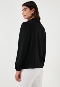 Black blouse with a structured collar, long puffed sleeves, and elastic cuffs, made from a smooth fabric. White pants partially visible.