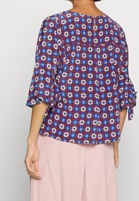 Blouse à motifs floraux avec des carrés multicolores en bleu, blanc et rouge. Dotée de manches trois-quarts et d'une ouverture en clé à l'arrière.