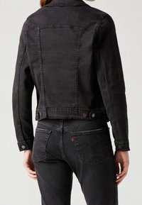 Chaqueta de mezclilla negra con un diseño recortado, que presenta cierres de botones y costuras visibles. Combinada con jeans de lavado oscuro y bolsillos traseros.