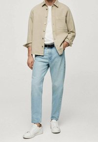 Camisa beige de botones, camiseta blanca, jeans de mezclilla azul claro y zapatillas blancas. Presenta un bolsillo frontal en la camisa, corte relajado y cinturón visible.