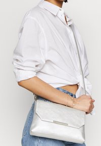 Stříbrná kovová crossbody taška s úhlovým designem, hladkým povrchem a výrazným kovovým akcentem. Nosit s bílou košilí a džínami.