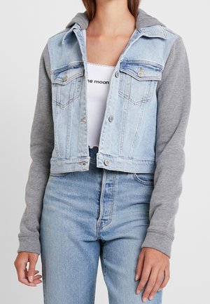Femme portant une veste en jean bleu clair avec des manches de sweat gris, associée à un jean bleu clair et un haut blanc avec un texte.
