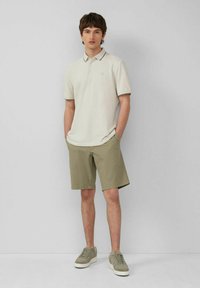 Beige Poloshirt mit Kragen und subtilen Besätzen, kombiniert mit hellen Khakihosen. Getragen mit grünen Turnschuhen, vor einem neutralen Hintergrund.