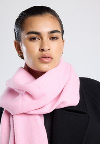 Sciarpa di lana rosa chiaro appoggiata attorno al collo, caratterizzata da una texture morbida e da una forma ampia e rettangolare, abbinata a un cappotto nero.