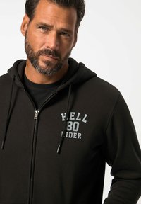 Felpa nera con zip e coulisse, con una stampa grafica grigia "HELL 80 RIDER" sul petto. Texture morbida, vestibilità casual e design con cappuccio.