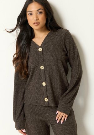 Femme aux longs cheveux foncés portant un cardigan en maille côtelée marron avec des boutons dorés et un pantalon assorti, debout contre un mur clair.