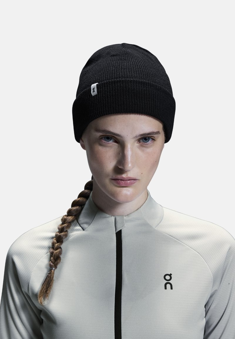 Un bonnet en tricot noir à côtes, arborant une petite étiquette logo blanche. Porté avec un haut gris clair ajusté, doté d'un col montant et d'un détail zippé.