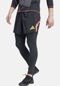Pantaloni sportivi neri con dettagli arancioni e un logo verde, sovrapposti a leggings neri aderenti e scarpe sportive nere con suole texturizzate.