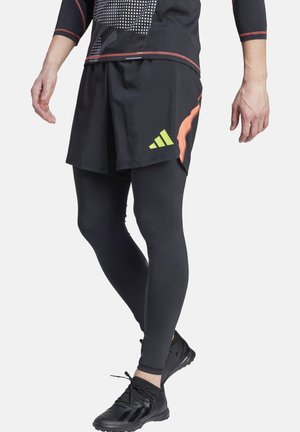 Shorts de sport noirs avec des accents orange et un logo vert, portés sur des leggings noirs ajustés et des chaussures de sport noires avec des semelles texturées.