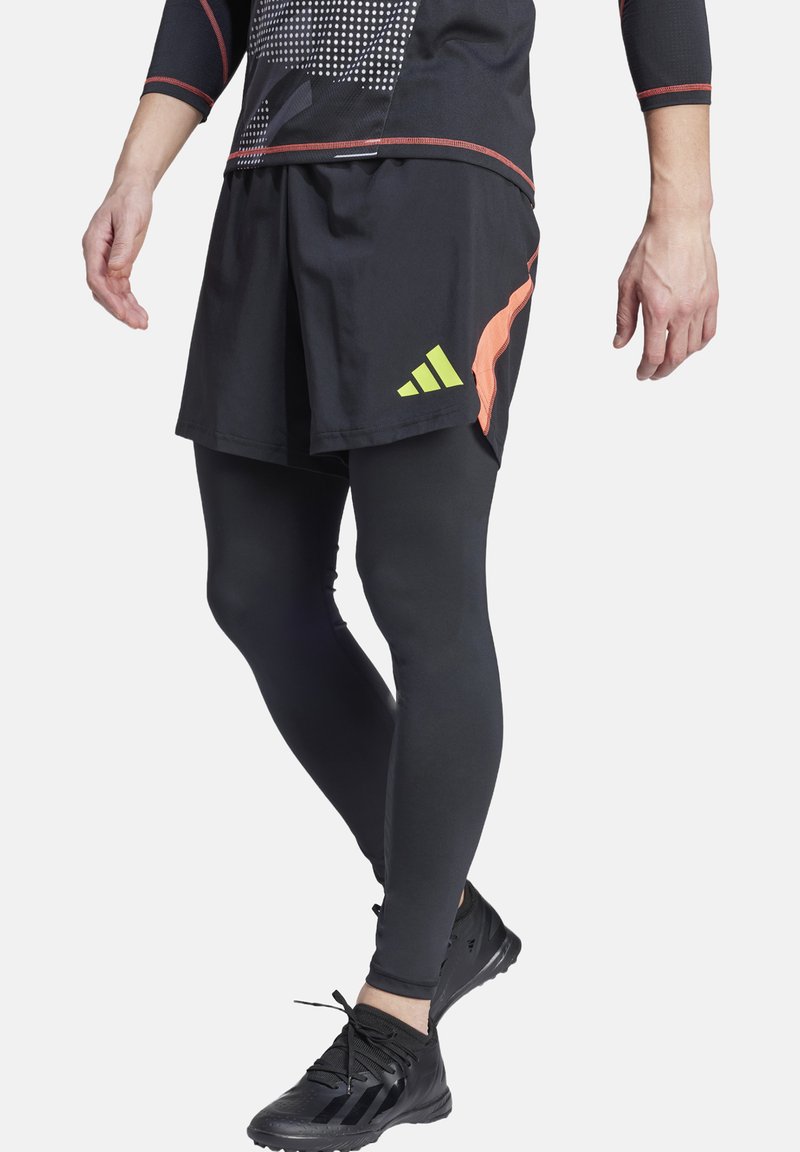Zwarte sportshorts met oranje accenten en een groen logo, over zwarte aansluitende leggings en zwarte sportschoenen met een gestructureerde zool.