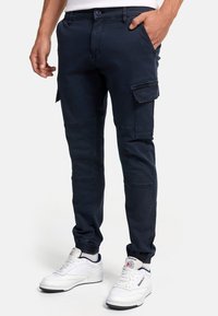 Homme portant un pantalon cargo bleu marine avec des poignets élastiques et des baskets blanches Reebok, debout avec une main dans la poche.
