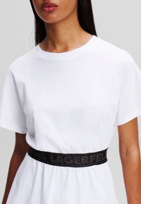 Bílé bavlněné tričko s krátkými rukávy, s černým elastickým páskem s nápisem "LAGERFELD". Jednoduchý, přiléhavý střih, hladká textura.