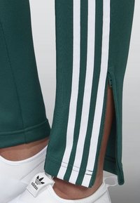 Zielone spodnie sportowe z białymi paskami po bokach i zamkiem błyskawicznym na dole. Wykonane z gładkiego, elastycznego materiału. Noszone z białymi butami.