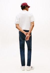 Mężczyzna w białej polo, ciemnych dżinsach, białych trampkach i czerwonej czapce stoi ze splecionymi za plecami rękami na tle jednolitego białego tła.