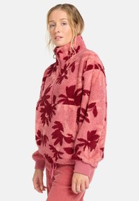 Fleece-Pullover in zartem Rosa mit dunkelroten Blattformen. Verfügt über einen hohen Kragen, einen halben Reißverschluss und elastische Bündchen, kombiniert mit pinken Hosen.