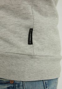 Primer plano del dobladillo de un sudadera gris claro con una etiqueta negra "CHASIN'" y unos jeans de denim azul visibles en la parte inferior.