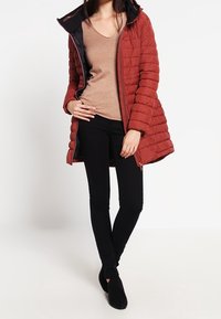 Röd quiltad pufferjacka med svart huva, dragkedjestängning och sidofickor; tillsammans med en beige topp och svarta leggings. Svarta ankelboots.