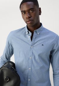 Blue and white checkered button-down shirt with a collar, featuring a small logo, held by a person carrying a black backpack.