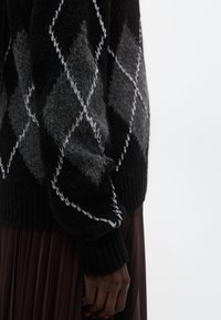 Maglione a motivo romboidale nero e grigio, texture a maglia, polsini a coste, abbinato a una gonna plissettata marrone scuro, vista laterale dei dettagli del tessuto.