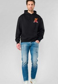 Zwarte hoodie met een voorste buidelzak, met een kleurrijke berenafbeelding; gecombineerd met lichtblauwe distressed jeans en witte sneakers.