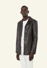 Bruine leren jas met een kraag, knoopsluiting en voorzakken. Draag deze over een wit shirt en combineer met lichtgekleurde broeken.