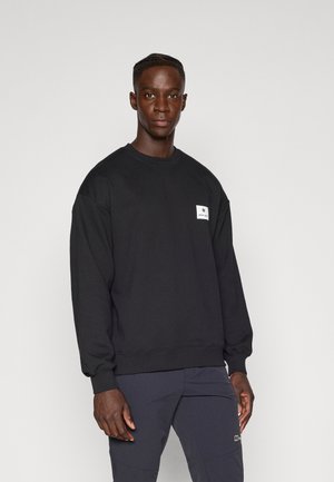 Sweatshirt noir avec un col rond, des poignets et un ourlet côtelés, et une petite étiquette blanche sur la poitrine. Tissu en coton doux, coupe décontractée.