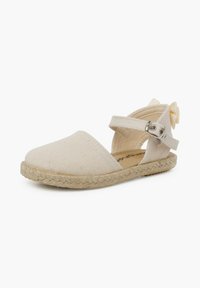 Beige canvas espadrille met een gevlochten jute zool, kruislings banden en een decoratieve strik bij de hiel. Plat ontwerp, gesloten neus.