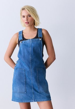 Blondynka ubrana w niebieską sukienkę ogrodniczkową z denimu i czarny top bez rękawów na jednolitym jasnym tle.