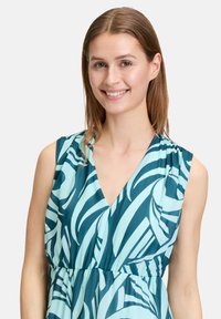 Mouwloze jurk met een diepe V-hals, gemaakt van vloeiende stof met turquoise en lichterblauwe bladpatronen, gathering in de taille.