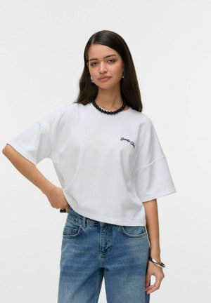 White oversized t-shirt with short sleeves featuring black embroidered text. Paired with light blue denim jeans and a black beaded necklace.