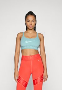 Žena stoji okrenuta prema naprijed, noseći svijetloplavi Nike sportski grudnjak i svijetlocrvene Nike Pro tajice, na jednostavnoj pozadini.
