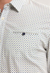 Camicia a maniche corte in tessuto bianco con un motivo a rombi blu e verdi. Presenta una tasca sul petto con bottone e un colletto con bottoni.