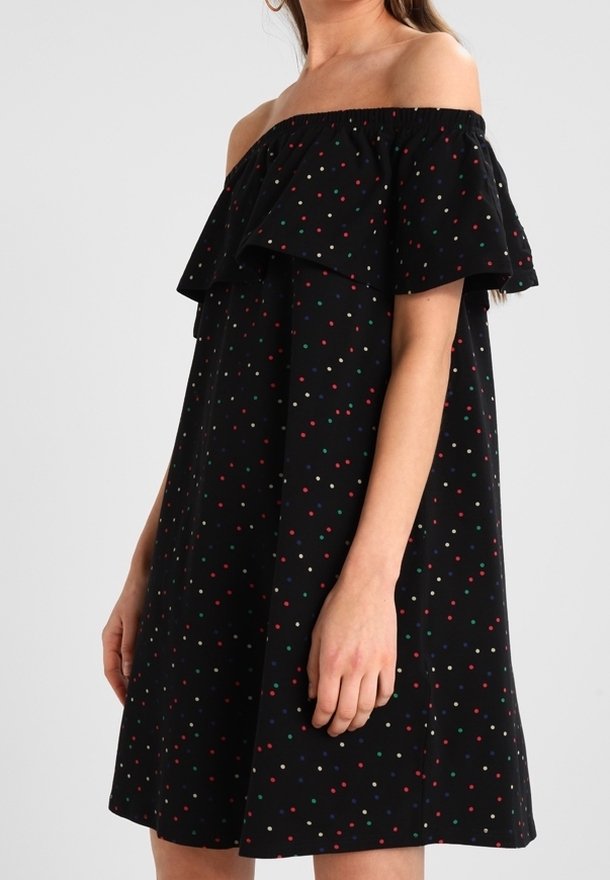 Femme portant une robe noire à épaules dénudées avec un col à volants et de petits pois multicolores, debout devant un fond uni.